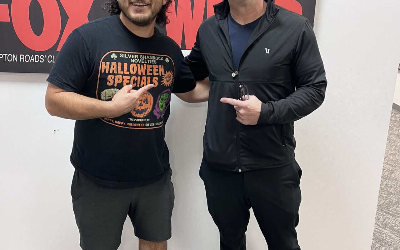 Two men pose indoors in front of radio station posters, smiling with thumbs up.