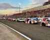 Race cars lined up on the track at sunset, with a crowded grandstand in the background and a fence separating pit area from spectators.