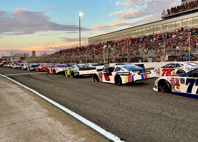 Race cars lined up on the track at sunset, with a crowded grandstand in the background and a fence separating pit area from spectators.