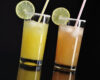 Two highball glasses with lime wheels and sugar rims, containing yellow and orange drinks with colorful straws on a dark background.