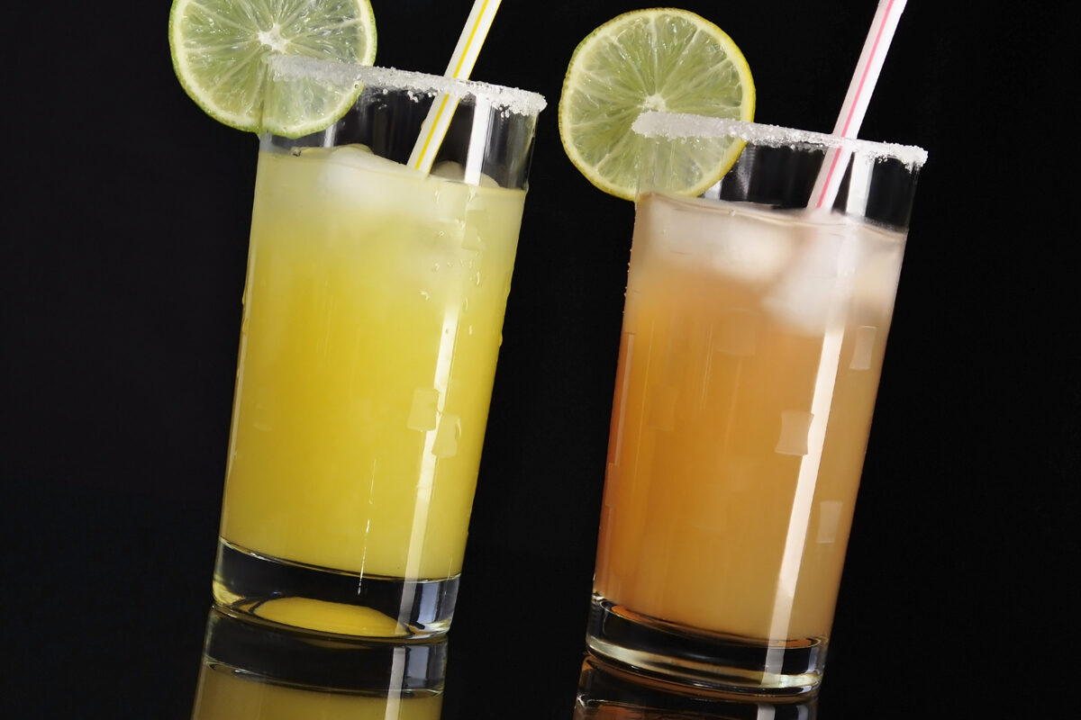 Two highball glasses with lime wheels and sugar rims, containing yellow and orange drinks with colorful straws on a dark background.