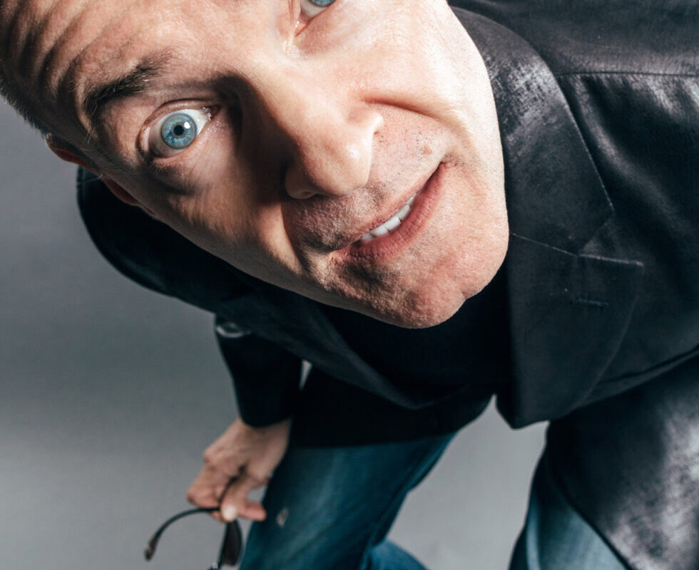 Man leaning toward the camera with a surprised expression, blue eyes wide, holding sunglasses in one hand.