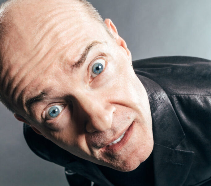 Man leaning toward the camera with a surprised expression, blue eyes wide, holding sunglasses in one hand.