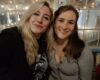 Two friends smiling at the camera in a cozy cafe with warm string lights and a glass wall in the background.
