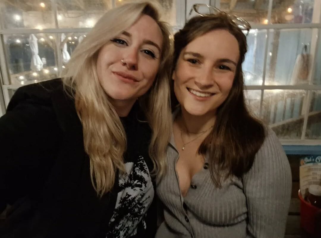 Two friends smiling at the camera in a cozy cafe with warm string lights and a glass wall in the background.