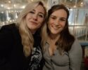 Two friends smiling at the camera in a cozy cafe with warm string lights and a glass wall in the background.