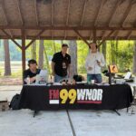 Radio hosts broadcasting from a shaded pavilion, three men at a table with mics, laptops, and equipment; banner reads FM 99 WNOR; outdoor park setting with trees in background.