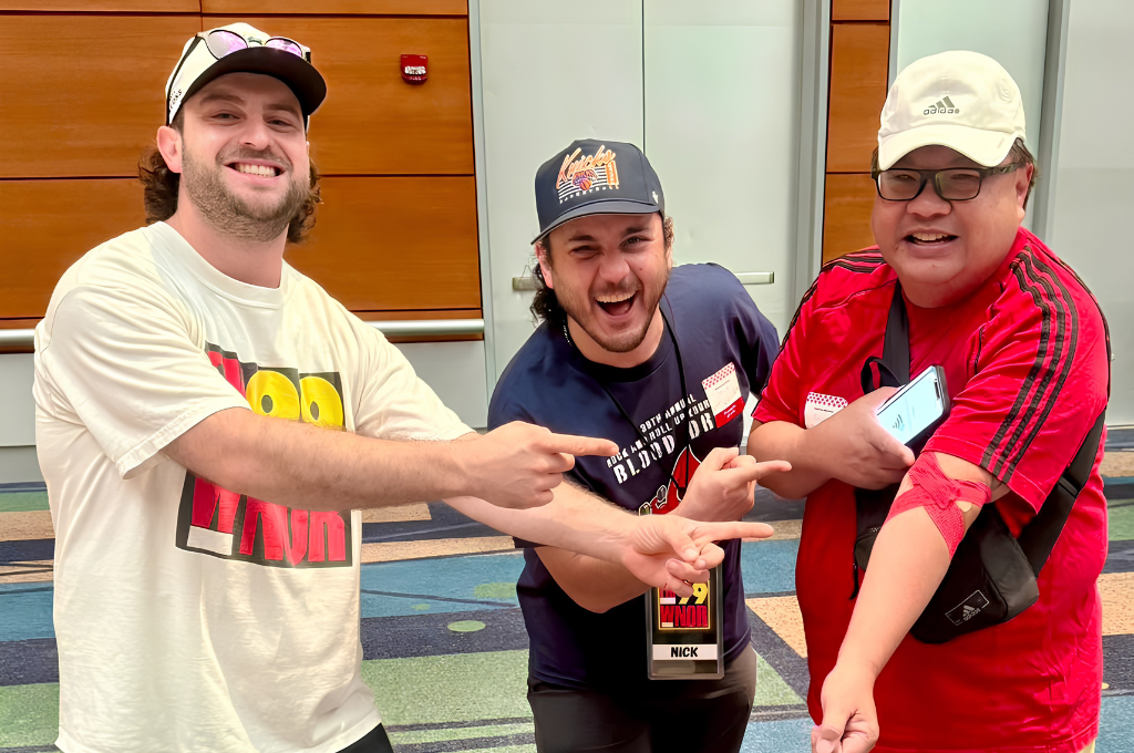 Three men in a hallway smiling and pointing at each other toward the camera.