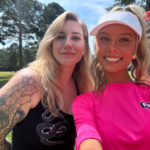 Two women taking a sunny park selfie; one has a full sleeve tattoo and wears black, the other wears a bright pink shirt and a white visor.
