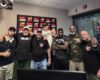 Eight men pose together in a radio studio, some with logo patches, arms around each other, with a 'Rumble Morning' poster in the background and radio gear nearby.