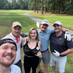 Five friends posing for a selfie on a sunny golf course with trees and a cart path in the background.
