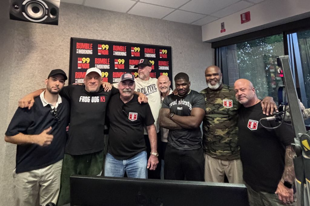 Eight men pose together in a radio studio, some with logo patches, arms around each other, with a 'Rumble Morning' poster in the background and radio gear nearby.