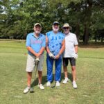 Three men posing for a photo on a golf course, each holding a golf club, with trees in the background.