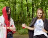 Two women in a forest toasting red drinks in clear plastic cups, smiling at the camera.