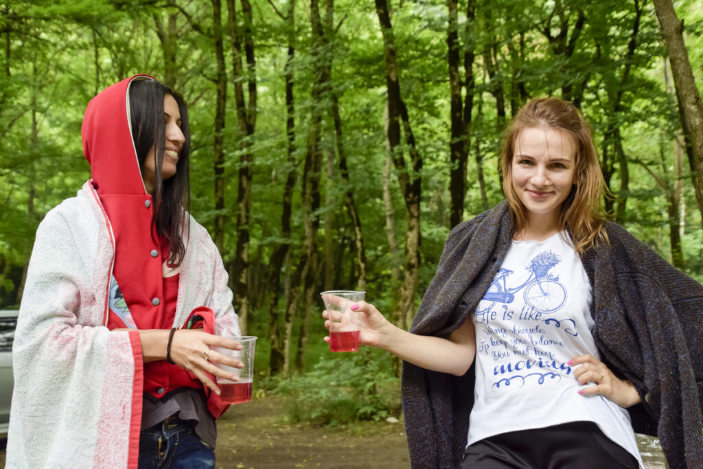 Two women in a forest toasting red drinks in clear plastic cups, smiling at the camera.