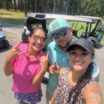Three friends posing for a selfie beside a golf cart on a sunny golf course, all smiling.