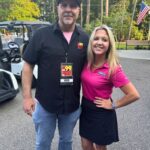 Two radio hosts posing outdoors at an event; man wears a Pinky's hat and badge labeled 'Rod', woman in a pink polo stands beside him with a wristwatch and sunglasses on her head.