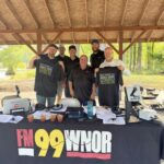Six men in matching black shirts pose behind a table with radios and laptops under a wooden pavilion at an outdoor broadcast setup.
