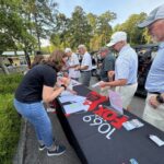 People sign in at an outdoor registration table beside golf carts, papers and badges on the table.