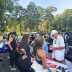 Group of people queuing at a registration table outdoors beside golf carts, with trees in the background and a clear sky.
