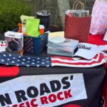 Outdoor vendor table with golf prizes and gear: Titleist cap, Bushnell box, Supersoft boxes, gift bag, and a patriotic star-spangled tablecloth.