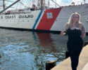 Woman in a black outfit standing on a dock beside a U.S. Coast Guard ship, smiling at the camera with the ship's red, white, and blue hull nearby.