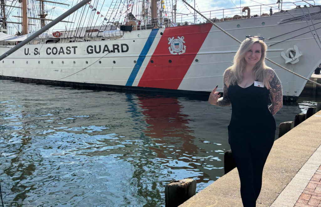 Woman in a black outfit standing on a dock beside a U.S. Coast Guard ship, smiling at the camera with the ship's red, white, and blue hull nearby.