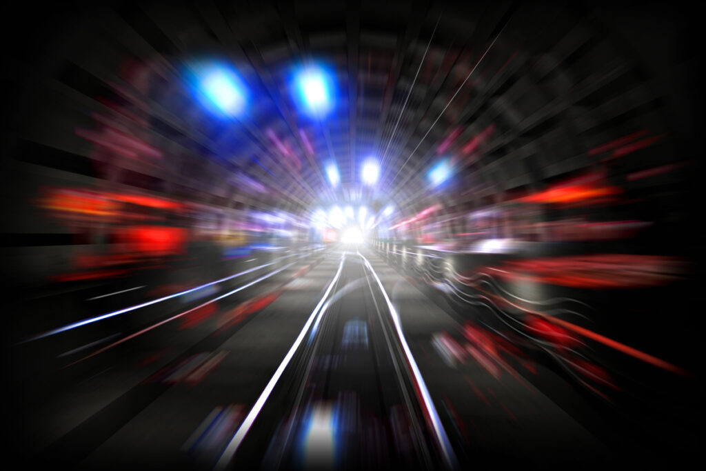 Blurred, long-exposure image of a tunnel with glowing blue and red lights streaking toward a bright center, conveying speed and motion.
