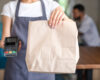 Server in a gray apron holds out a brown paper bag and a card payment terminal for a takeout order