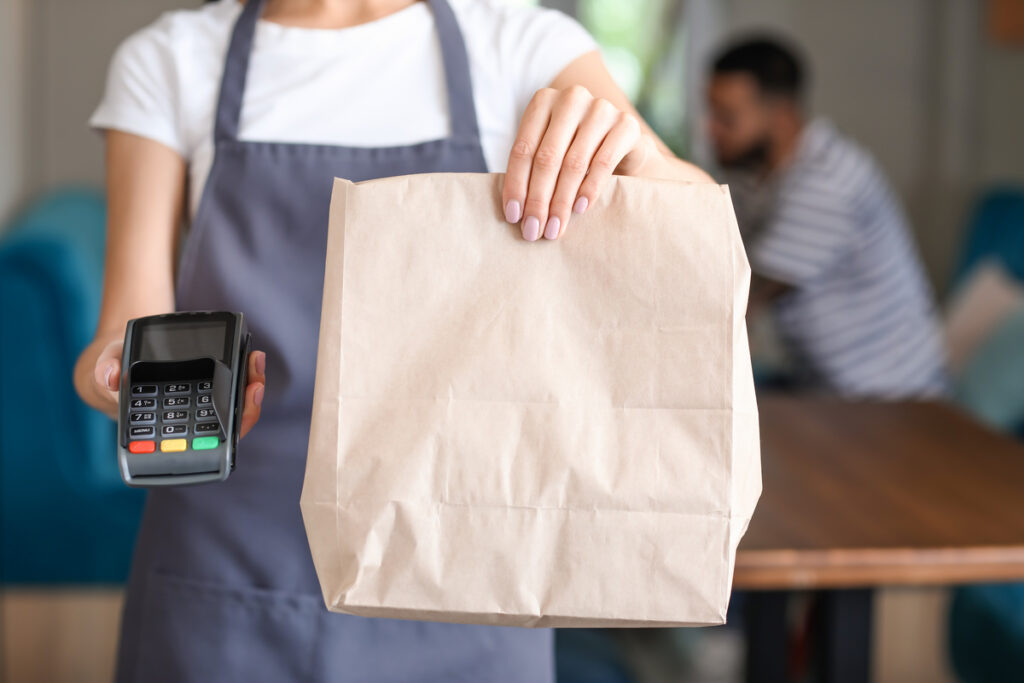 Server in a gray apron holds out a brown paper bag and a card payment terminal for a takeout order