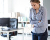 Woman in a blue striped blouse stands in an office, arms crossed over her midsection and a worried expression.