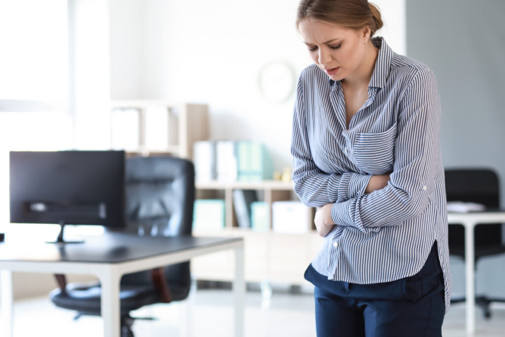 Woman in a blue striped blouse stands in an office, arms crossed over her midsection and a worried expression.