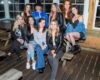 Nine young women posing for a group photo on a wooden patio, smiling at the camera. (informative, group portrait)