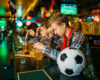 Group of friends at a sports bar with beers and plates of food, a soccer ball on the bar foreground.