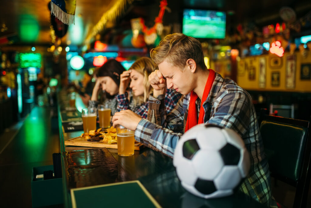 Group of friends at a sports bar with beers and plates of food, a soccer ball on the bar foreground.